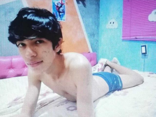 Tulipanti, sem camisa, com cabelo preto, deitado em uma cama com roupa íntima azul, de bruços, olhando para a câmera, em um quarto com cores vibrantes..