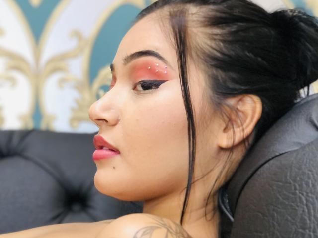 MadelineEgan's face with pink eye makeup, black eyeliner, red lips, and black hair in a messy bun, leaning on a black leather chair. She has a tattoo on her shoulder.