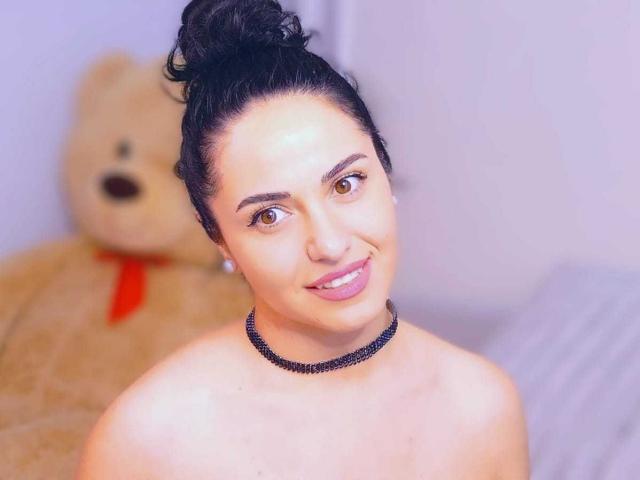 Jenniferwild69 with black hair in a bun, brown eyes, pink lipstick, black choker, smiling, showing bare shoulders, and a large beige teddy bear with a red ribbon in the background.