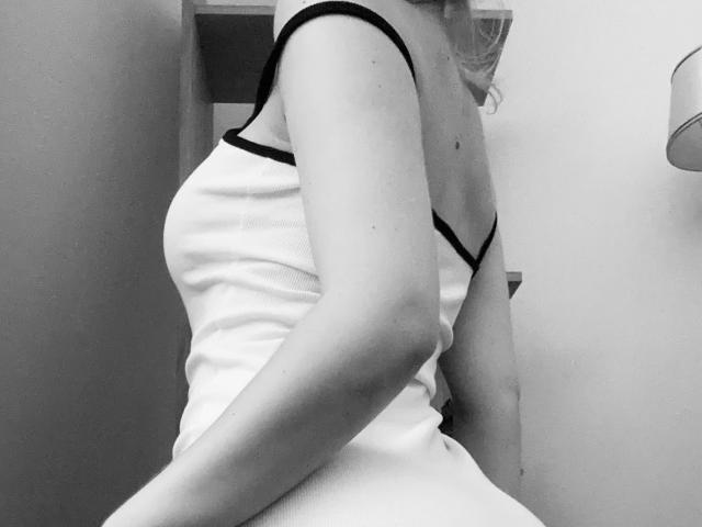 SugarrX in a white tank top with black straps, side profile, showing her tits and arm, in a black-and-white photo, seated with a slight twist.