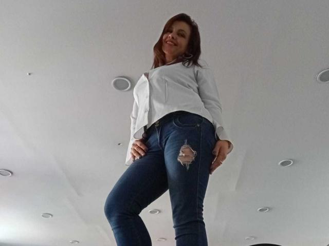 MilenaHott69 with long brown hair, white shirt, and ripped blue jeans, standing with hands on hips, looking down at camera, smiling. Low angle shot.