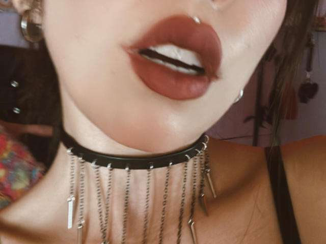 Het gezicht van IzzysMisss, met de focus op haar glanzende bruine lippen en een neusring, draagt een zwarte choker met metalen kettingen en een zwarte band. Haar huid is licht, haar lippen zijn vol..