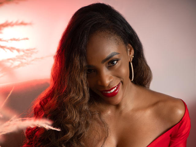 IsabellaKaned has dark brown skin, wavy brown hair, red lipstick, smiling, wearing a red off-shoulder top, and gold hoop earrings, with red lighting on her left side.