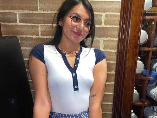 AlessiaFerry with long black hair, glasses, and a white and blue button-up shirt, showing her small tits and a tattoo on her left arm, smiling.