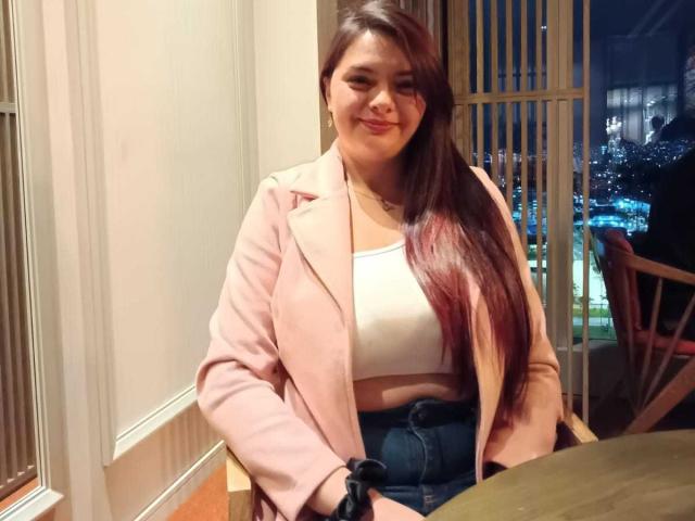 KatherinLatin, with long brown hair, pink blazer, white crop top, and blue jeans, smiling at the camera, has a cute face and small tits, sitting at a table.