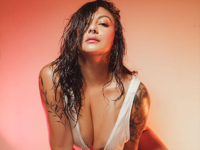 Karlaparadise with wet, dark hair, nose ring, and white tank top, showing large tits and tattooed arm, leaning forward with a sultry expression. Warm red-orange lighting.