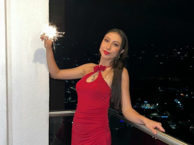 Emma Wells, com cabelos castanhos longos, vestindo um vestido vermelho, segurando uma faísca, em pé na varanda à noite, com batom vermelho, parede branca, luzes da cidade ao fundo, braço direito apoiado na grade, braço esquerdo levantado..