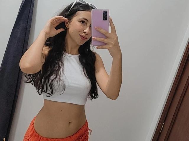 Emma Wells, com cabelos longos e pretos, blusa branca cropped que mostra a cintura e shorts laranja, tirando uma selfie no espelho com um celular rosa, sorrindo e usando óculos de sol brancos..