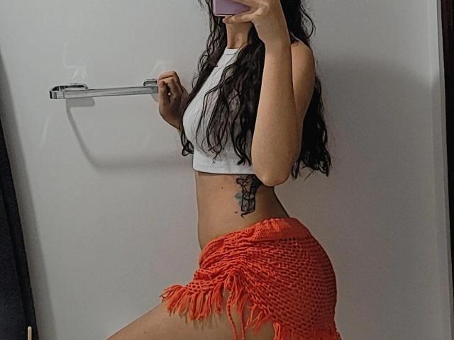 Emma Wells, con el cabello negro y mojado, un top blanco corto y una falda naranja con flecos, sosteniendo un teléfono, mostrando un tatuaje en el lado izquierdo, con un trasero pequeño y visible en un selfie en el espejo..