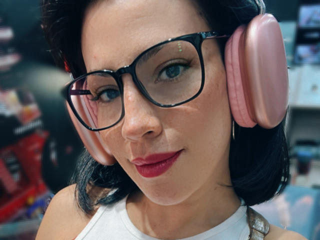 Liliysweet, met roze over-ear hoofdtelefoons, een zwarte bril, rode lippenstift en een witte top, heeft een lichte huid, blauwe ogen en zwart haar dat tot op haar schouders reikt, en kijkt recht in de camera..