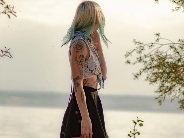 AmazingSugar-hot with blonde and blue-green hair, sleeve tattoos, tied-up shirt, black skirt, standing by water, hand near face, soft sunlight, gentle breeze, calm atmosphere.