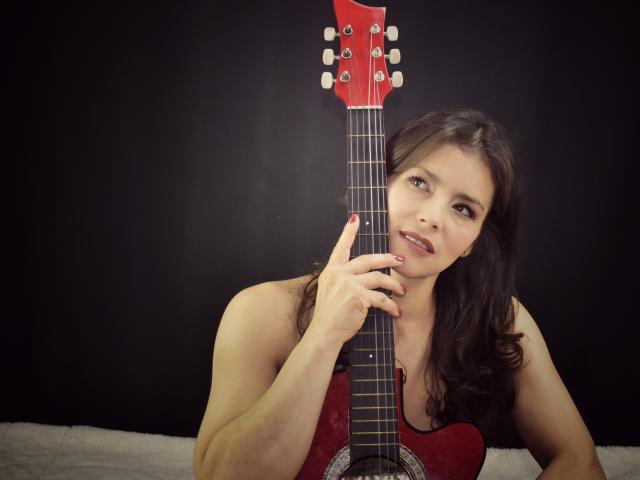 Scottie with brown hair, holding a red electric guitar, her hand on the neck, smiling, looking slightly to the side, black background, white bed, visible tits.
