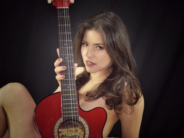 Scottie with long brown hair, holding a red acoustic guitar, showing off her tits, sitting with knees up, black background, red nails, slight smile, soft lighting.