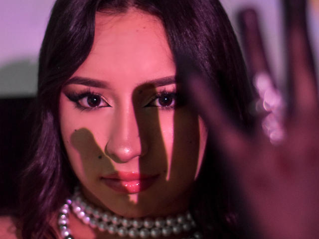 MiaSuarez has dark purple hair, heavy eyeliner, and glossy pink lips. She's wearing a pearl necklace, with shadows from a hand in the foreground on her face.