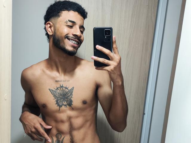 AfroJake, zonder shirt, met een getatoeëerde borst en buikspieren, die een telefoon vasthoudt, lachend, met kort krullend haar, een baard en een kleine neusring, staand in een spiegel voor een selfie..
