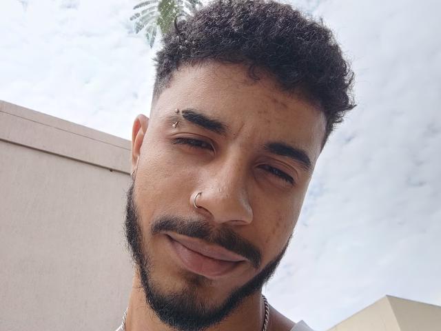 AfroJake has curly black hair, a mustache, beard, nose ring, and a small stud in his right eyebrow. He's smiling slightly with his eyes half-closed. Sky and building in background.