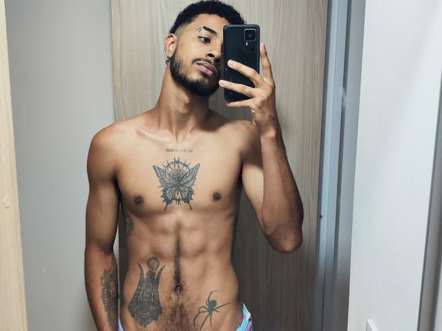 AfroJake, torse nu, tenant un téléphone, la langue tirée, tatouages sur la poitrine et le bas du ventre, musclé, peau foncée, cheveux courts et bouclés, barbe, portant des sous-vêtements colorés, autoportrait pris dans un miroir..