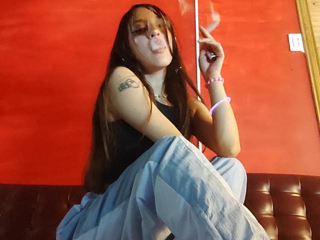 CamilaMarie, met lang bruin haar, die rookt, een zwarte tanktop draagt, een blauwe rok, een roze armband, en een sigaret vasthoudt, heeft een tatoeage op haar rechterarm en zit op een bruine bank..