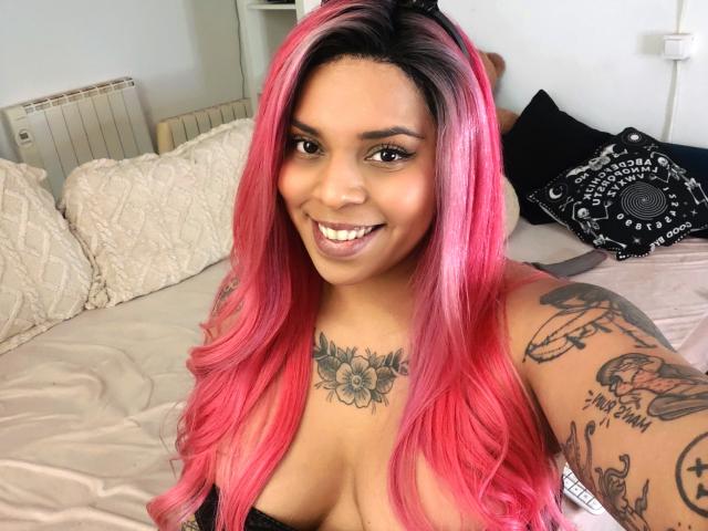 Chili met felroze haar, grote tieten en tatoeages lacht en laat haar borst- en armtatoeages zien, liggend op een bed.