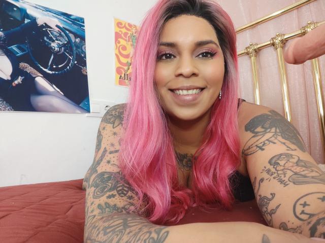 Chili with pink hair and tattoos is smiling, lying on a bed, her hand near an erect penis in the background.