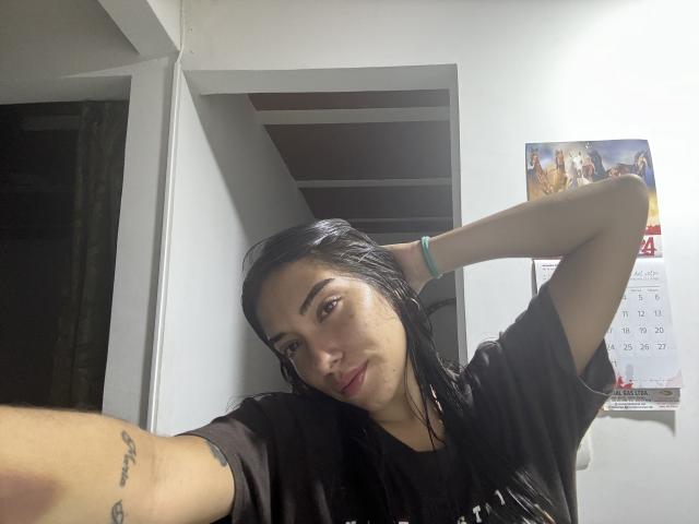 SharonKlart with black hair, tan skin, and a tattoo on her arm, wearing a black shirt, looking at the camera with a slight smile, hand behind head.