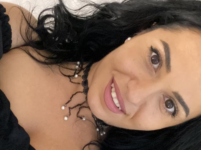 Jenniferwild69 with black curly hair, brown eyes, and pink lipstick, smiling, wearing a black lace choker with pearl beads, showing her tits, and a black off-shoulder top.