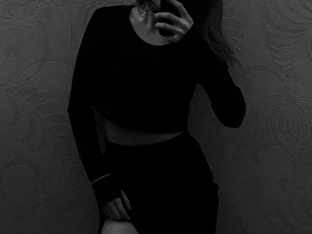 AlexSantorski in a black long-sleeve crop top and black skirt, holding a phone to her face, with a hand on her thigh, in a dark, grainy photo.
