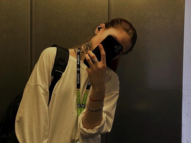 AlexSantorski taking a selfie, wearing a white shirt, black backpack, and a green lanyard, with earbuds in, phone covering face, tattoo visible on neck, in dim light.