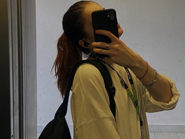 AlexSantorski with red ponytail, white shirt, black backpack, holding phone covering face, ring on left hand, bracelet on right wrist, taking mirror selfie, showing phone camera.