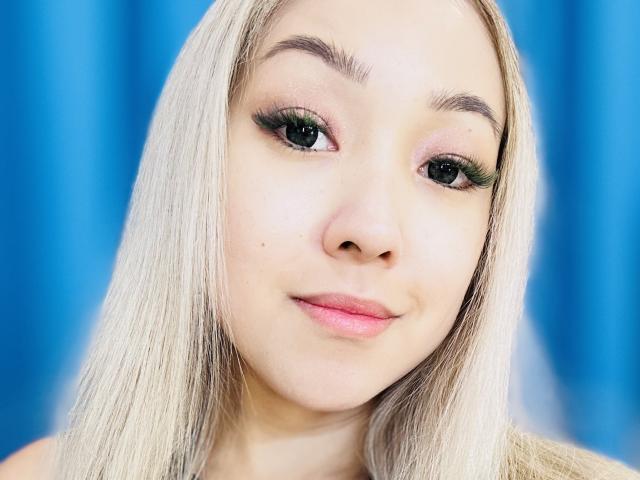RenyLime has blonde hair, green eyeshadow, long green eyelashes, pink lips, and a small beauty mark on her left cheek. She's looking at the camera with a slight smile.