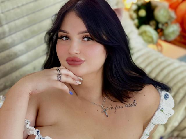 MonaSteele with black hair, green eyes, and pink lipstick, showing off her tits, wearing a white off-shoulder top, hand on chin, blue nail, and tattoo on chest.