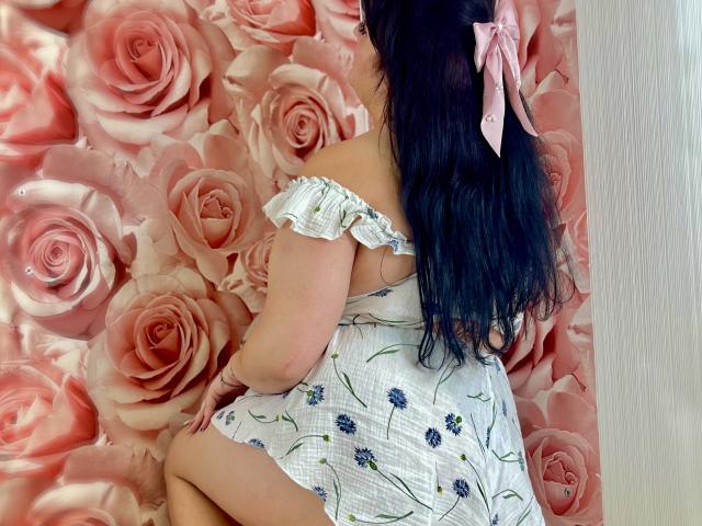 MonaSteele with long black hair, pink bow, off-shoulder white dress with blue flowers, kneeling, showing her ass, against pink rose wallpaper.