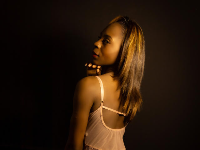 Angeiiina with medium brown skin, looking over her shoulder, wearing a sheer pink top, her brown hair with blonde highlights, holding a string of small lights.