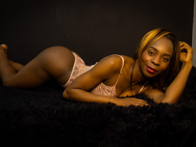 Angeiiina, laying on a black furry surface, wearing pink lace lingerie, with her ass up, looking at the camera, smiling with red lipstick, and blonde highlights in her hair.