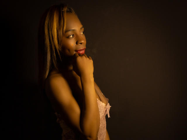 Angeiiina with dark skin, blonde highlights, wearing pink lace lingerie, biting her red-lipped finger, looking to the side, in dim light, showing her small tits and slim figure.
