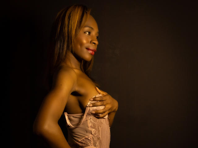 Angeiiina with dark skin, blonde highlights, and red lipstick, covering her small tits with pink lace lingerie, smiling in low light, dark background.