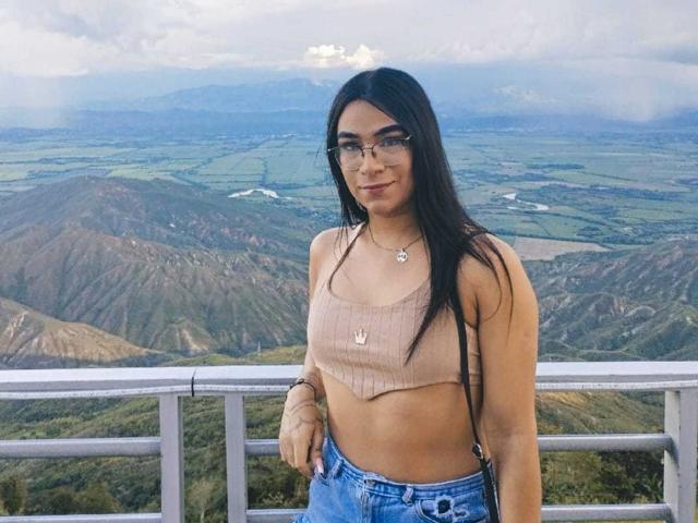 MiaMarinHot with long black hair, glasses, tan crop top showing her tits, and blue jeans, standing in front of a mountain view, smiling slightly.