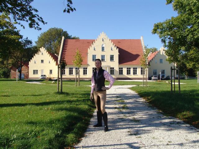 AuroraVanLaakFetish-hot stands on a gravel path, wearing a pink shirt, black vest, brown pants, and black boots, in front of a yellow, red-roofed, medieval-style house.
