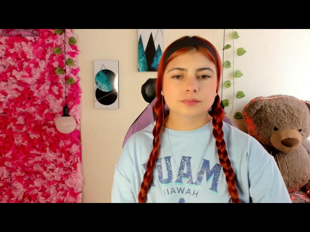 Isabella69s with red braids in a blue "UCLA" shirt, standing in front of a pink leafy wall, small blue and green mountain paintings, and a brown teddy bear.