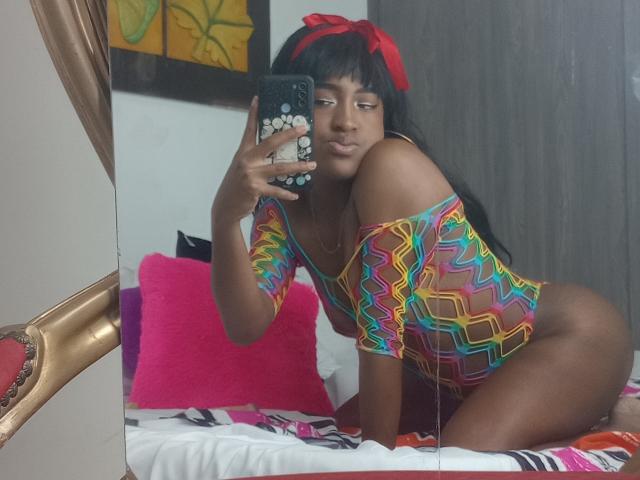 LilaJamez with dark skin, black hair, red bow, and colorful zigzag top, kneeling on bed, taking mirror selfie, showing ass, looking at phone with white flower case.