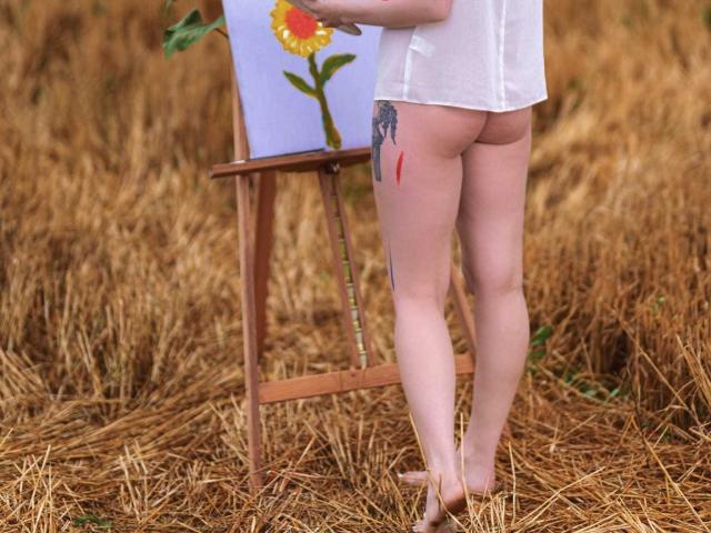 LoreenBlanc stands bare-assed in front of an easel with a sunflower painting, wearing a white shirt, showing a tattoo on her right thigh, in a field of dry grass.