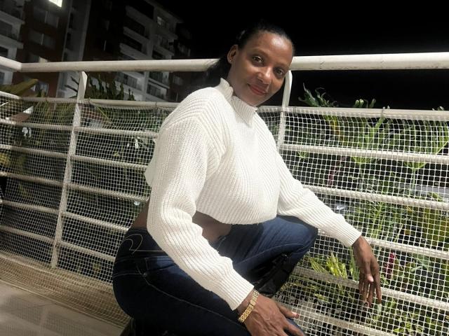 Anahismaar with dark skin, crouching, wearing a white knit sweater, exposing a slim waist, and tight blue jeans, smiling, with a black bracelet, on a rooftop at night.