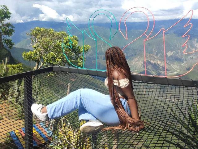 AkaneCox with long brown braids, sitting on a black mesh fence, wearing a white off-shoulder top and light blue jeans, facing mountains, colorful neon outlines in the background.