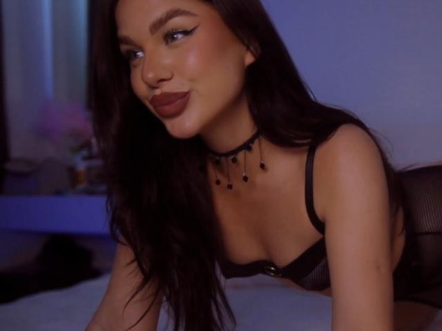 JaninaPussy with long dark hair, wearing black lingerie and a black choker with beads, looking to the side with a slight smile, showing her tits and a cute nose.