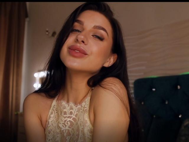 JaninaPussy with long dark hair, lace top, and a slight smirk, looking down with a cute, relaxed expression. Her tits are covered, but her pretty face is visible.