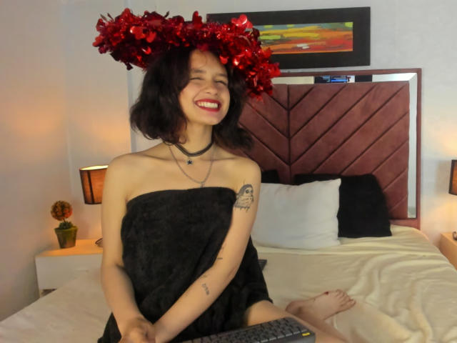 CatarsisNous com uma peça de cabeça floral vermelha, toalha preta sem alças cobrindo os seios, sorrindo, sentada na cama, tatuagem no braço esquerdo, colar, batom vermelho, pés descalços, cama com travesseiros brancos e pretos..