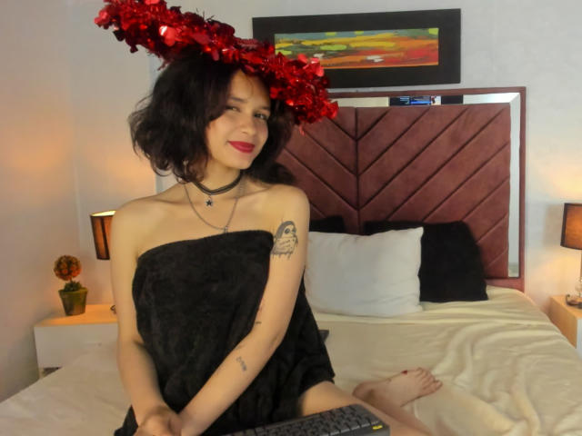 CatarsisNous mit dunklen, lockigen Haaren, einem roten Blumen-Haarschmuck, einem schwarzen Handtuch, das Brüste und Gesäß bedeckt, einem Tattoo am rechten Arm, rotem Lippenstift, sitzend auf einem Bett, lächelnd, mit einer Choker-Kette..