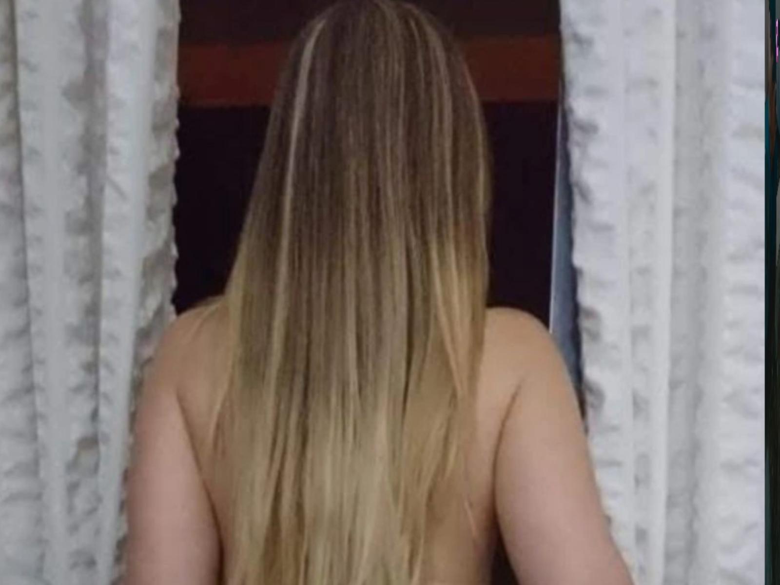 LeticiaSpinerx's bare back with long, blonde, straight hair, facing a window, framed by white curtains. Her upper body is visible, topless.