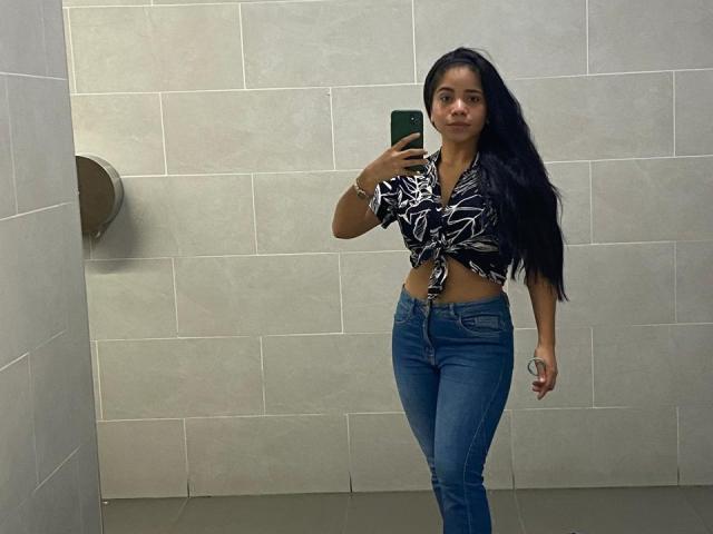 SaraSweetx, avec de longs cheveux noirs, porte une chemise noire et blanche à motifs de feuilles, un jean bleu, tient un téléphone vert, se tient dans la salle de bain, montre ses fesses et son ventre, et regarde le miroir..