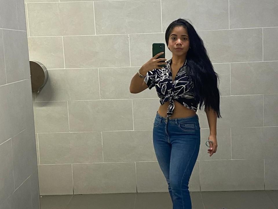 SaraSweetx, avec de longs cheveux noirs, porte une chemise noire et blanche à motifs de feuilles, un jean bleu, tient un téléphone vert, se tient dans la salle de bain, montre ses fesses et son ventre, et regarde le miroir..
